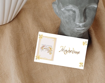 Könnte beinhalten: Eine weiße Karte mit dem Namen "Madeline" in goldener Schrift und eine kleinere Karte mit floralen Illustrationen und dem Text "Gratitude is the bridge to abundance." Die Karten liegen auf einer beigen Oberfläche, in der Nähe einer Steinbüste.