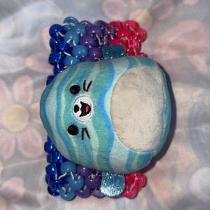 May include: A plush, blue and white striped creature with a smiling face is attached to a beaded bracelet. The bracelet features blue, purple, and pink beads. The plush has a white interior.