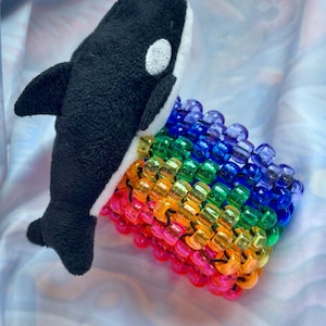 Op de afbeelding: Een zacht speelgoed orka in zwart en wit, naast een regenboogkleurige kralenarmband. De armband is gemaakt van transparante kralen in een spectrum van kleuren: roze, oranje, geel, groen, blauw en paars.