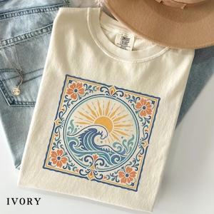 May include: Ivory-colored t-shirt featuring a circular design with a sun, wave, and floral elements in orange, blue, and yellow. The shirt is made of a soft material and is paired with blue jeans and a necklace.