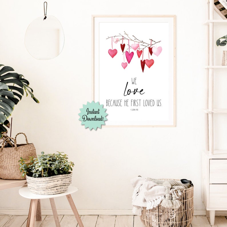 May include: A printable wall art print with a watercolor illustration of pink and red hearts hanging from a branch. The text reads "We love because He first loved us. - 1 John 4:19"