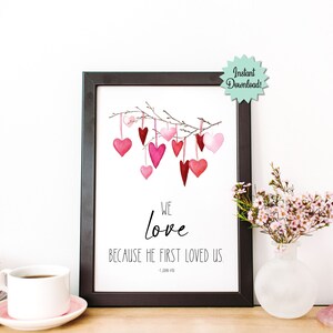 May include: A printable wall art print with a watercolor illustration of pink and red hearts hanging from a branch. The text reads "We love because He first loved us. - 1 John 4:19"