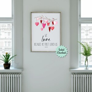 May include: Printable wall art with a watercolor illustration of a branch with pink and red hearts hanging from it. The text reads "We love because He first loved us - 1 John 4:19".