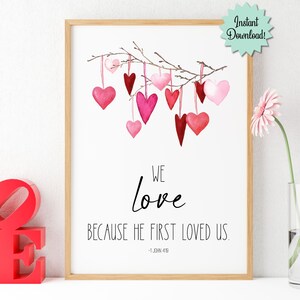 May include: Printable wall art featuring a watercolor illustration of pink and red hearts hanging from a branch. The text reads "We love because He first loved us. - 1 John 4:19"