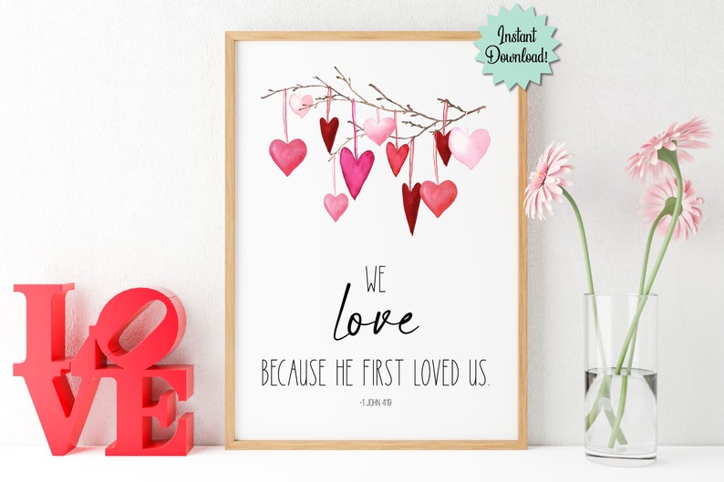 May include: Printable wall art featuring a watercolor illustration of pink and red hearts hanging from a branch. The text reads "We love because He first loved us. - 1 John 4:19"