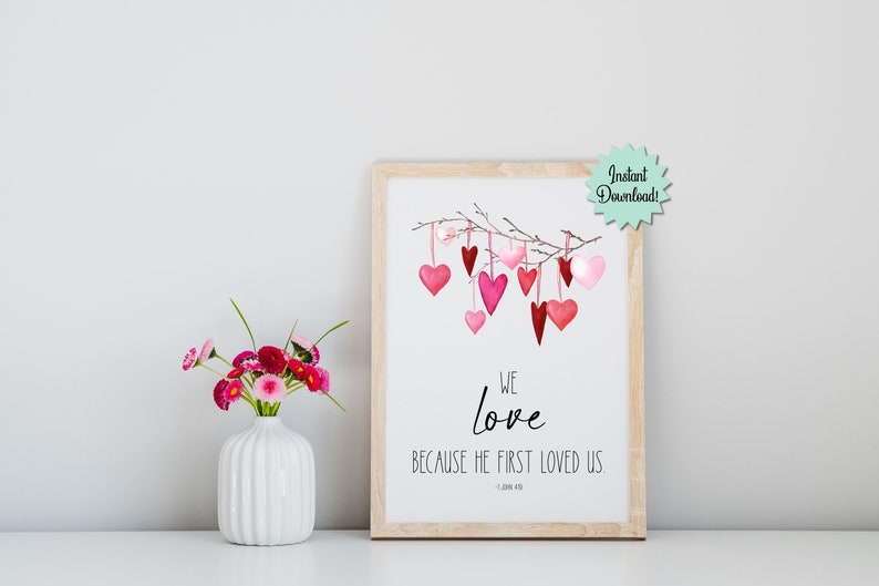 May include: Printable wall art with a watercolor illustration of pink and red hearts hanging from a branch. The text reads "We love because He first loved us - 1 John 4:19".