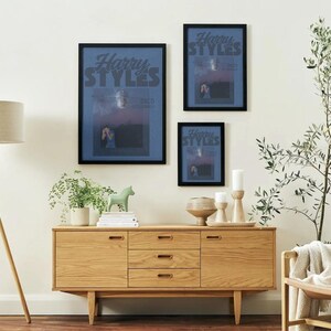 Può includere: Tre poster incorniciati con la scritta "Harry Styles" sono appesi a una parete bianca. Un mobile in legno con cassetti e una lampada con paralume beige sono di fronte al muro. Un cavallo decorativo verde e delle piante sono sul mobile.