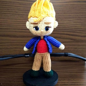 Puede incluir: Muñeco de ganchillo hecho a mano de un niño con pelo amarillo, ojos azules y una chaqueta azul sobre una camisa roja. El muñeco tiene pantalones color canela y zapatos verdes.