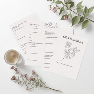 May include: A flat lay featuring printed paper sheets with text, including "CEO Action Plan" and "CEO Time Block." The sheets are accompanied by a small cup and sprigs of flowers, all arranged on a white surface. The text is in black.