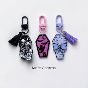 May include: Three coffin-shaped keychains with lobster clasps and tassels. The black one features skeletons and flowers, the pink one shows a ghost and bats, and the light purple one displays a bow, spiderweb, and the text R.I.P. More Charms.