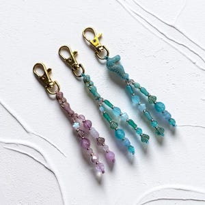 May include: Three keychains featuring gold-tone lobster clasps and dangling beaded strands. One keychain has purple beads, while two feature blue and green beads, including seahorse-shaped charms and various small shell and star-shaped accents.