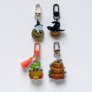 May include: Four acrylic keychains featuring Halloween themes. The top left shows a cat in a pumpkin with a gold clasp. The top right is a pumpkin wearing a witch hat with a black clasp. The bottom left has a green smoke pumpkin and orange tassel with an orange clasp. The bottom right features a stack of three pumpkins with a silver clasp.