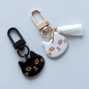 May include: Two cat-shaped keychains featuring glossy faces with gold-colored line details. One keychain has a black cat charm with a black lobster clasp, while the other has a white cat charm, a gold-colored lobster clasp, and a white tassel.