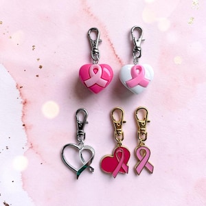May include: Five metal lobster clasp charms featuring breast cancer awareness themes. Two charms are heart-shaped with pink ribbons, one is a silver open heart, one is a gold heart with a pink ribbon, and one is a gold pink ribbon charm.