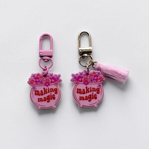 May include: Two keychains featuring a pink cauldron design with red text that reads making magic and a floral arrangement on top. One keychain has a pink lobster clasp, while the other has a gold lobster clasp and a small pink fabric tassel.