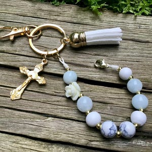 Puede incluir: Un llavero dorado con un dije de cruz, una borla blanca y una pulsera con cuentas azules y blancas. El llavero tiene un mosquet&oacute;n y una anilla partida. La pulsera presenta una peque&ntilde;a flor blanca y detalles dorados.