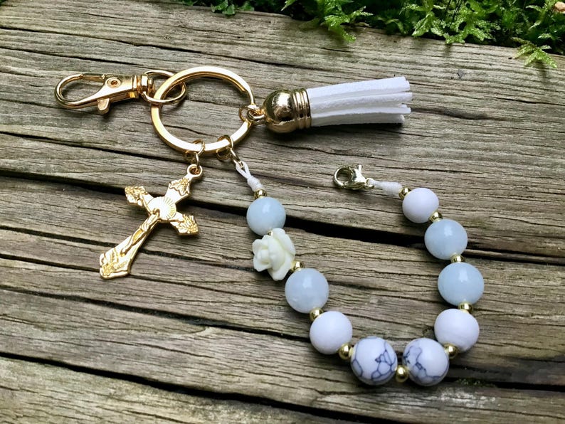 Puede incluir: Un llavero dorado con un dije de cruz, una borla blanca y una pulsera con cuentas azules y blancas. El llavero tiene un mosquet&oacute;n y una anilla partida. La pulsera presenta una peque&ntilde;a flor blanca y detalles dorados.