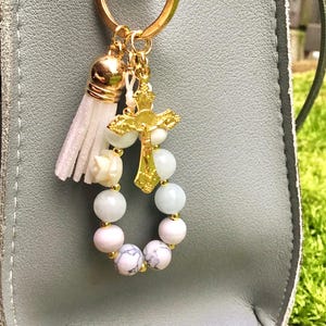 Puede incluir: Un llavero dorado con un colgante de cruz y una peque&ntilde;a borla. El llavero presenta un anillo de cuentas blancas y azules. El colgante de cruz est&aacute; detallado con una figura de Jes&uacute;s. La borla es blanca y las cuentas son redondas.