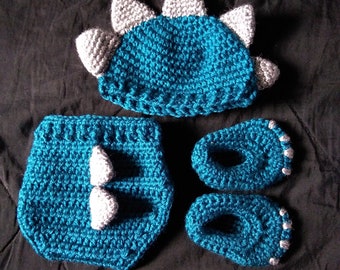May include: A crocheted dinosaur baby outfit in teal blue and gray. The outfit includes a hat, diaper cover, and booties.