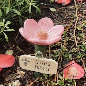 May include: A pink flower-shaped garden stake with a textured center and a green stem. A small wooden sign with a bee illustration reads "CUPS FOR BEE". The stake is surrounded by greenery and fallen petals.