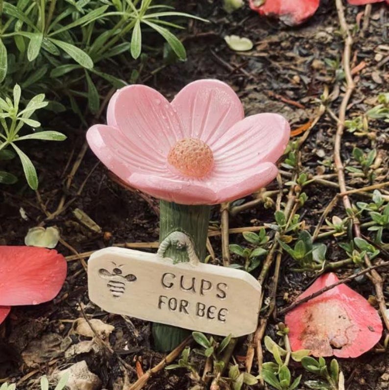 May include: A pink flower-shaped garden stake with a textured center and a green stem. A small wooden sign with a bee illustration reads "CUPS FOR BEE". The stake is surrounded by greenery and fallen petals.