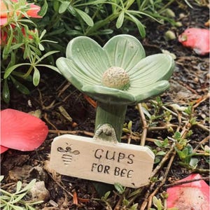 May include: A green ceramic flower-shaped garden stake with a beige center and a small sign that reads "CUPS FOR BEE" with a bee illustration. The stake is surrounded by greenery and red flower petals.