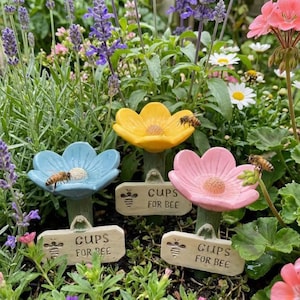 May include: Three ceramic flower-shaped bee water cups in a garden setting. The flowers are yellow, blue, and pink, each with a bee perched on them. Small signs read "CUPS FOR BEE". The garden has lavender and other flowers.