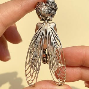 May include: A silver-toned pendant in the shape of a fairy or insect. The pendant features detailed wings with an intricate webbed design. The fairy's head is adorned with a crown of leaves. The pendant is held between two fingers.