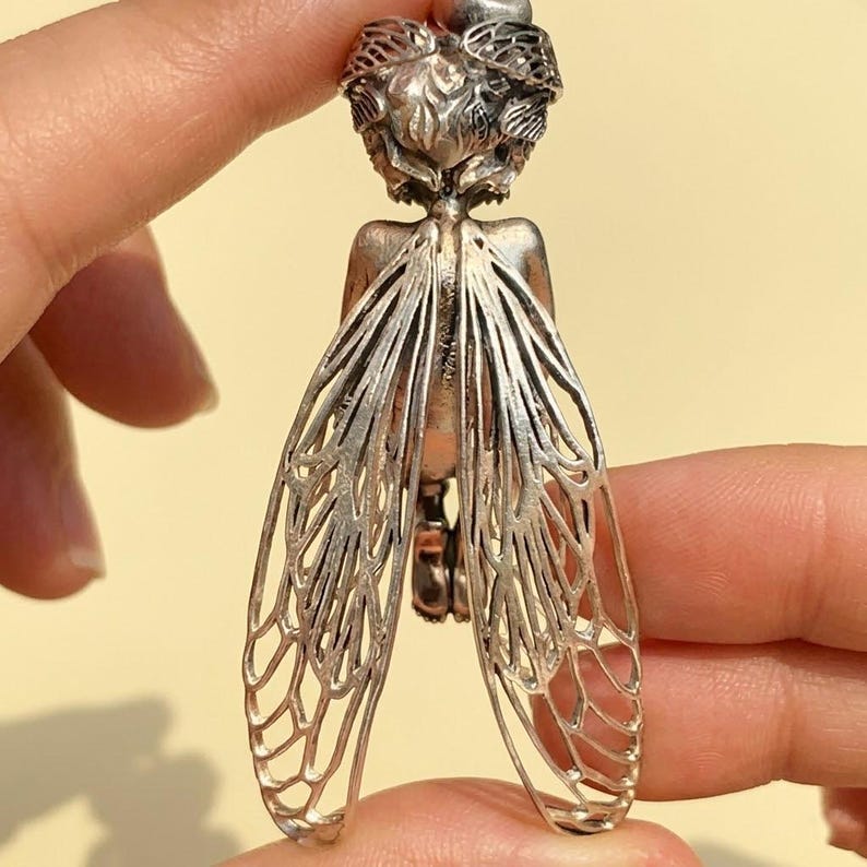 May include: A silver-toned pendant in the shape of a fairy or insect. The pendant features detailed wings with an intricate webbed design. The fairy's head is adorned with a crown of leaves. The pendant is held between two fingers.