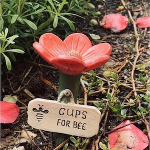 May include: A coral-colored flower-shaped garden ornament with a green stem. A small wooden sign reads "CUPS FOR BEE" with a bee illustration. The ornament is set in a garden bed with greenery and fallen petals.