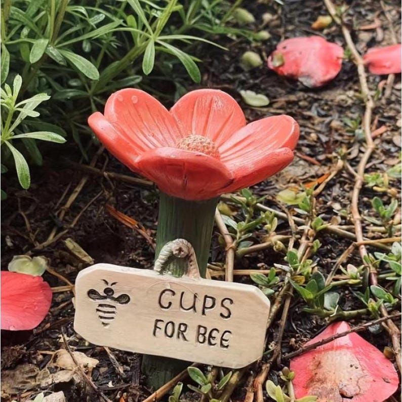 May include: A coral-colored flower-shaped garden ornament with a green stem. A small wooden sign reads "CUPS FOR BEE" with a bee illustration. The ornament is set in a garden bed with greenery and fallen petals.