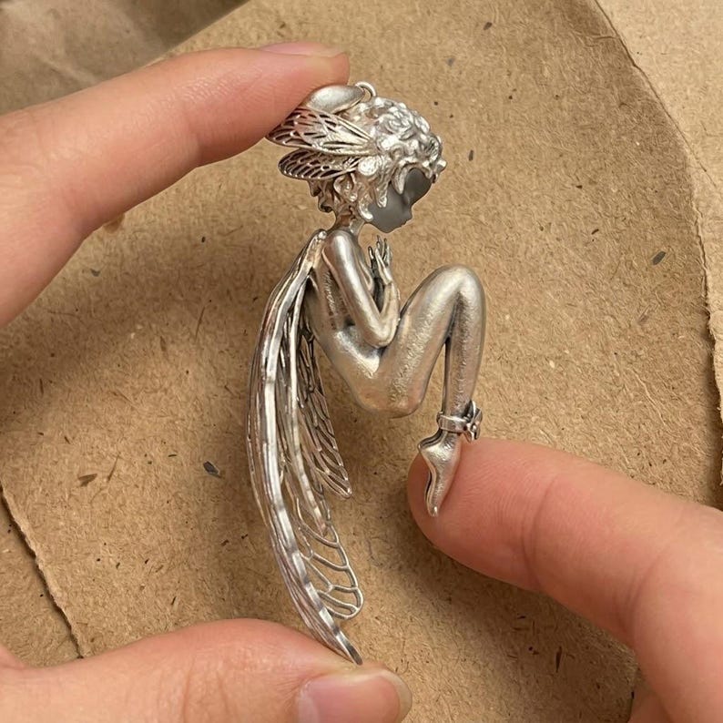 May include: A silver-toned fairy figurine with detailed wings, hair, and a seated pose. The fairy is holding its hands near its face. The wings are intricately designed. The figurine is held between two fingers.