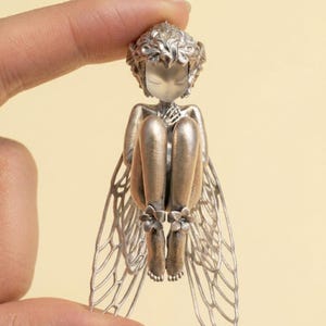 May include: A silver-toned fairy figurine with delicate, detailed wings and curled hair. The fairy is seated with its knees drawn up, and its feet are adorned with small flower accents. The figurine is held between two fingers.