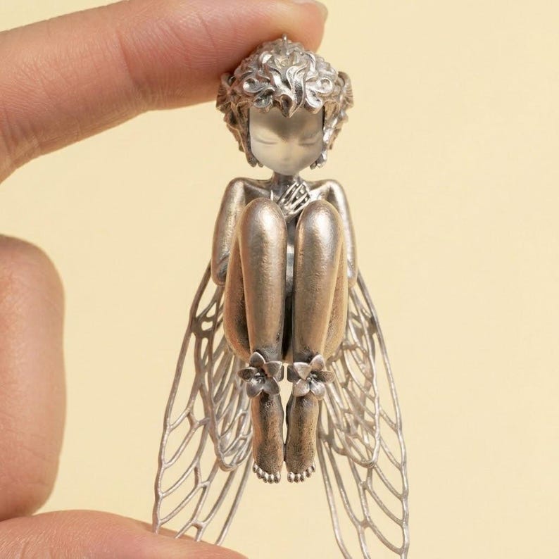 May include: A silver-toned fairy figurine with delicate, detailed wings and curled hair. The fairy is seated with its knees drawn up, and its feet are adorned with small flower accents. The figurine is held between two fingers.