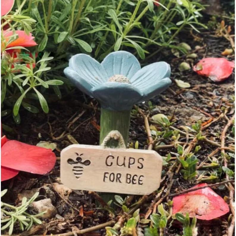 May include: A light blue flower-shaped garden ornament with a green stem and a small wooden sign that reads "CUPS FOR BEE". The sign features a bee illustration. The ornament is set in a garden with green plants and red petals.
