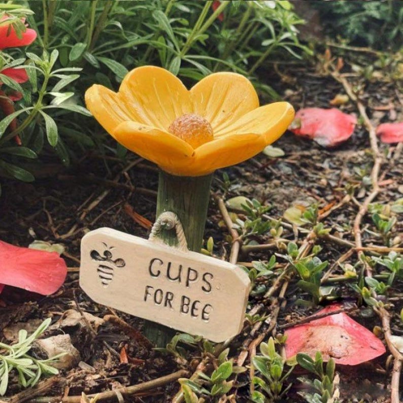 May include: A yellow flower-shaped garden ornament with a brown center and a green stem. A small sign reads "CUPS FOR BEE" with a bee illustration. The ornament is set in a garden with green plants and red petals.