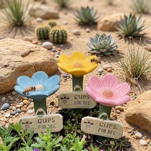 May include: Three flower-shaped bee water feeders in yellow, blue, and pink. Each has a sign that reads "Cups for Bee". Bees are present on the feeders. The feeders are in a garden setting with rocks and plants.
