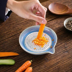 Puede incluir: Un rallador de cerámica con borde azul y superficie blanca se utiliza para rallar una zanahoria. Los trozos de zanahoria rallada son visibles en el rallador. Una cuchara de madera y un cuenco pequeño con granos de pimienta están en el fondo.