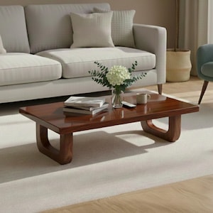 Könnte beinhalten: Ein rechteckiger, dunkelbrauner Couchtisch mit abgerundeten Beinen. Vor einer hellgrauen Couch mit weißen Kissen. Auf dem Tisch stehen eine Vase mit weißen Blumen, Bücher und eine Tasse. Der Tisch ist etwa 120 cm lang.