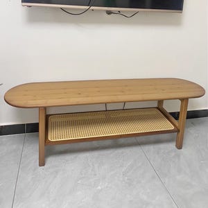 May include: A light brown wooden bench with an oval top and a lower shelf with a woven rattan design. The bench has four sturdy legs and is suitable for a living room or entryway.
