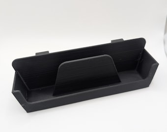 May include: A black, 3D-printed desk organizer with a rectangular base and a central divider. The organizer has a sloped front and two raised sections on the back. The interior is divided into three sections.