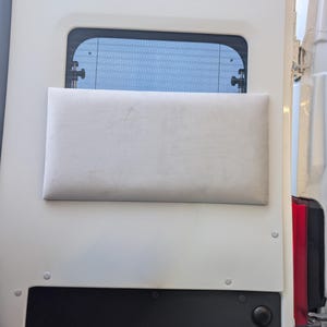 Może przedstawiać: Jasnoszara prostokątna poduszka zamontowana na białych drzwiach pojazdu, poniżej prostokątnego okna. Drzwi mają czarny panel na dole i są zabezpieczone widocznymi nitami.