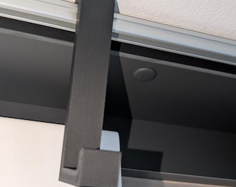 May include: A dark gray, L-shaped bracket, likely for a sliding door or panel, is mounted to a silver track. The bracket is made of a solid, matte material and is positioned against a white and black background.