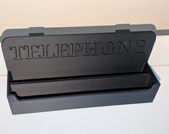 May include: A black, rectangular telephone holder with the word "TELEPHONE" embossed on the lid. The lid is open, revealing a compartment for storage. The holder is made of a matte material and sits on a white surface.