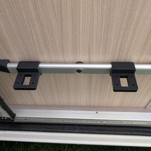 Puede incluir: Una barra de metal plateado con tapas de plástico negro y soportes de montaje, fijada a una superficie clara con vetas de madera. Los soportes tienen aberturas cuadradas. La barra es parte del interior de un vehículo.