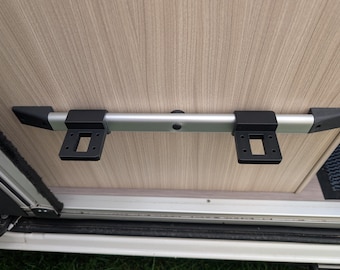 May include: A silver metal bar with black plastic end caps and mounting brackets, attached to a light wood-grain surface. The brackets have square openings for secure attachment. The bar appears to be part of a vehicle's interior.