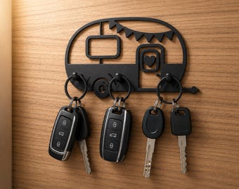 May include: A black metal wall-mounted key holder shaped like a vintage camper trailer. The design features a rectangular window, a door with a heart cutout, and decorative bunting. Three hooks hold four sets of car keys and metal house keys.