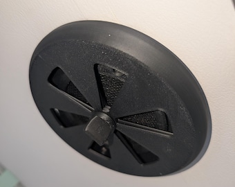 May include: A black, circular vent cover with a star-shaped design. The cover is mounted on a white surface and has a central, square-shaped fastener. The vent cover is made of a matte material.
