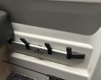 May include: A silver-colored horizontal wall-mounted rail featuring three black plastic hooks. The rail is attached to a light gray interior panel, with a small black plastic component visible on the left side of the metal bar structure.