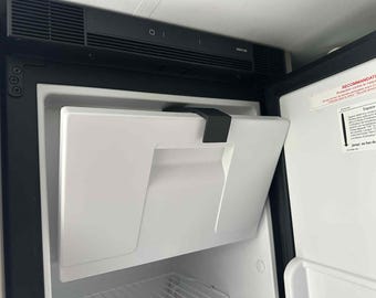 May include: A white refrigerator door is open, revealing the interior. The door has a handle and a black latch. The top of the refrigerator is black with a control panel.